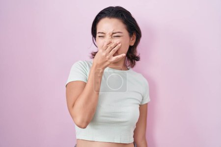 Téléchargez les photos : Jeune femme hispanique debout sur fond rose sentant quelque chose de puant et dégoûtant, odeur intolérable, retenant souffle avec les doigts sur le nez. mauvaise odeur - en image libre de droit