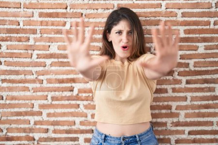 Téléchargez les photos : Jeune femme brune debout sur un mur de briques faisant un geste d'arrêt avec les mains paumes de la main, expression de colère et de frustration - en image libre de droit