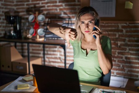 Photo for Young beautiful woman working at the office at night speaking on the phone looking unhappy and angry showing rejection and negative with thumbs down gesture. bad expression. - Royalty Free Image