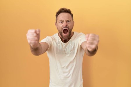 Photo for Middle age man with beard standing over yellow background angry and mad raising fists frustrated and furious while shouting with anger. rage and aggressive concept. - Royalty Free Image