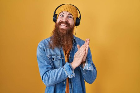 Téléchargez les photos : Homme caucasien à longue barbe écoutant de la musique à l'aide d'écouteurs applaudissant et applaudissant heureux et joyeux, souriant mains fières ensemble - en image libre de droit