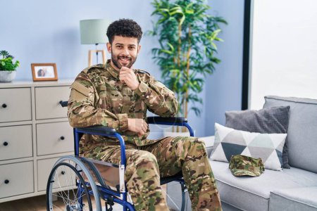 Foto de Hombre árabe vistiendo uniforme camuflaje del ejército sentado en silla de ruedas mirando confiado a la cámara sonriendo con los brazos cruzados y la mano levantada en la barbilla. pensar positivamente. - Imagen libre de derechos