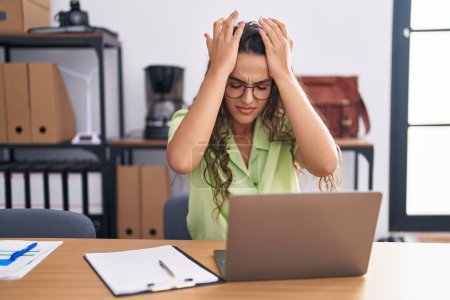 Jeune femme hispanique travaillant au bureau portant des lunettes souffrant de maux de tête désespérés et stressés parce que la douleur et la migraine. mains sur la tête. 