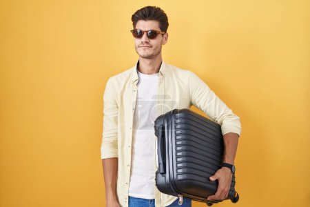 Téléchargez les photos : Jeune homme hispanique tenant une valise allant en vacances d'été souriant regardant vers le côté et regardant loin en pensant. - en image libre de droit