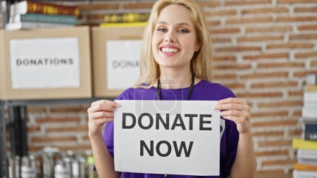 Téléchargez les photos : Jeune femme blonde bénévole souriant confiant tenant faire un don maintenant bannière au centre de charité - en image libre de droit
