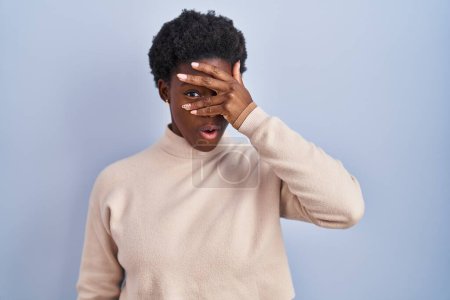 Foto de African american woman standing over blue background peeking in shock covering face and eyes with hand, looking through fingers with embarrassed expression. - Imagen libre de derechos