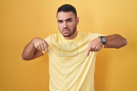 Foto de Young hispanic man standing over yellow background pointing down looking sad and upset, indicating direction with fingers, unhappy and depressed. - Imagen libre de derechos