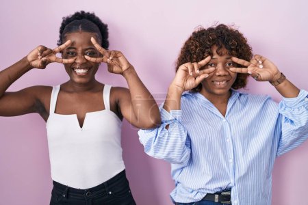 Photo for Two african women standing over pink background doing peace symbol with fingers over face, smiling cheerful showing victory - Royalty Free Image