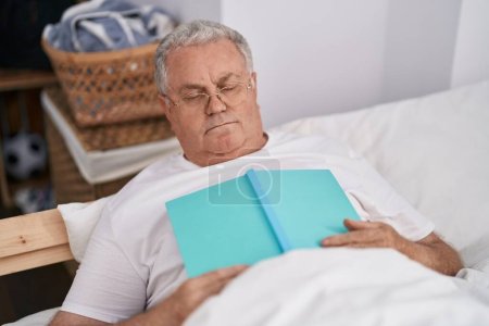 Téléchargez les photos : Moyen-âge homme aux cheveux gris couché sur le lit dormant tenant livre dans la chambre - en image libre de droit