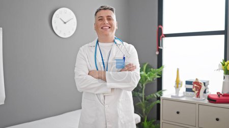 Foto de Joven caucásico hombre médico sonriendo confiado de pie con los brazos cruzados gesto en la clínica - Imagen libre de derechos