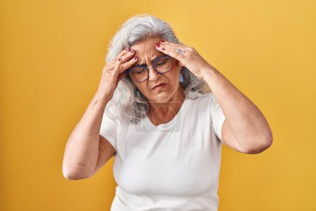 Photo for Middle age woman with grey hair standing over yellow background with hand on head for pain in head because stress. suffering migraine. - Royalty Free Image