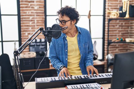 Foto de Young hispanic man musician singing song playing piano keyboard at music studio - Imagen libre de derechos