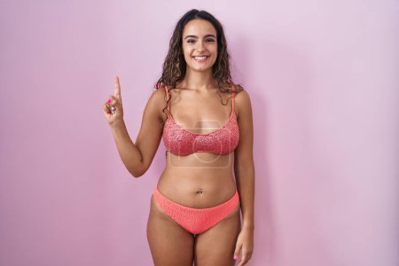 Téléchargez les photos : Jeune femme hispanique portant lingerie sur fond rose montrant et pointant vers le haut avec le doigt numéro un tout en souriant confiant et heureux. - en image libre de droit