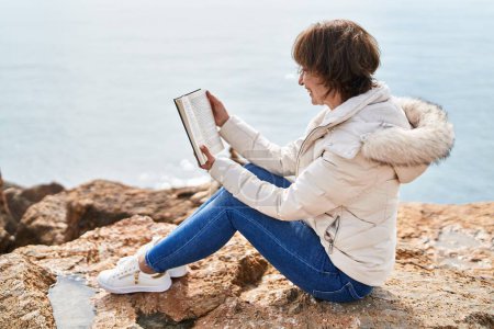 Téléchargez les photos : Middle age woman reading book sitting on the rock at seaside - en image libre de droit
