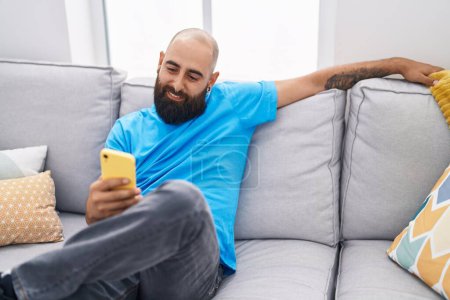 Téléchargez les photos : Jeune chauve utilisant smartphone assis sur le canapé à la maison - en image libre de droit