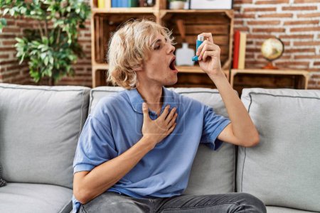 Photo for Young blond man using inhaler sitting on sofa at home - Royalty Free Image