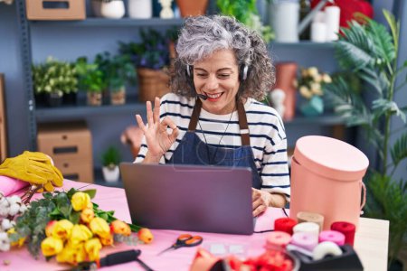 Téléchargez les photos : Femme d'âge moyen avec les cheveux gris travaillant chez le fleuriste avec ordinateur portable faisant signe ok avec les doigts, sourire geste amical excellent symbole - en image libre de droit