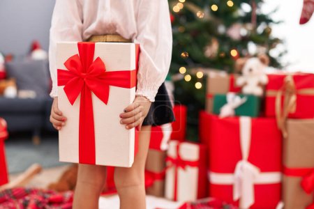 Foto de Adorable chica hispana sosteniendo regalo en la espalda de pie junto al árbol de Navidad en casa - Imagen libre de derechos