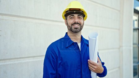 Photo for Young hispanic man worker wearing hardhat holding blueprints at construction place - Royalty Free Image