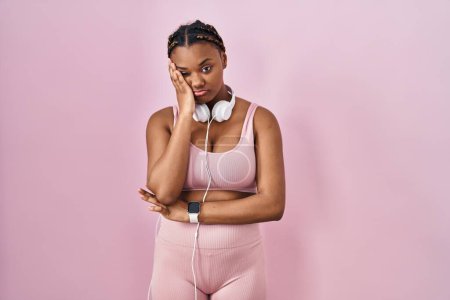 Téléchargez les photos : Femme afro-américaine avec des tresses portant des vêtements de sport et des écouteurs pensant fatigué et ennuyé avec des problèmes de dépression avec les bras croisés. - en image libre de droit