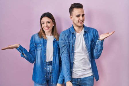 Foto de Joven pareja hispana de pie sobre fondo rosa sonriente alegre presentando y señalando con la palma de la mano mirando a la cámara. - Imagen libre de derechos