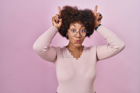 Téléchargez les photos : Jeune femme afro-américaine debout sur fond rose faisant un geste drôle avec le doigt sur la tête comme des cornes de taureau - en image libre de droit