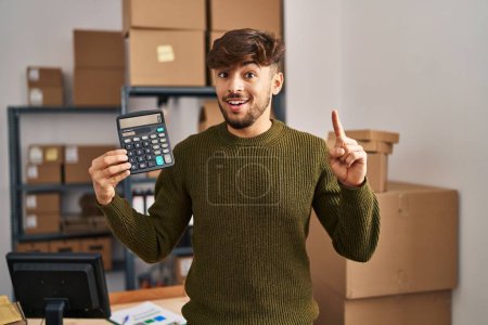 Photo for Arab man with beard working at small business ecommerce holding calculator surprised with an idea or question pointing finger with happy face, number one - Royalty Free Image