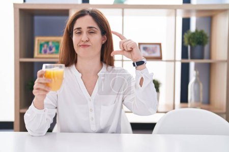 Téléchargez les photos : Femme brune buvant un verre de jus d'orange geste souriant et confiant avec la main faisant signe de petite taille avec les doigts regardant et la caméra. concept de mesure. - en image libre de droit