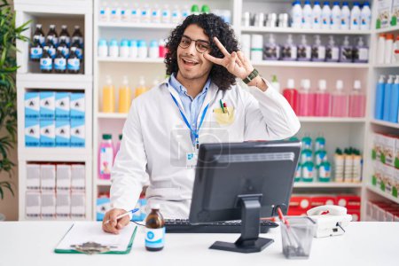 Téléchargez les photos : Homme hispanique aux cheveux bouclés travaillant à la pharmacie pharmacie faisant symbole de paix avec les doigts sur le visage, souriant joyeux montrant la victoire - en image libre de droit