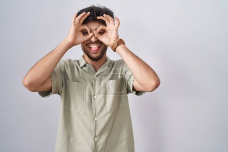 Foto de Hombre árabe con barba de pie sobre fondo blanco haciendo buen gesto como binoculares sacando la lengua, ojos mirando a través de los dedos. expresión loca. - Imagen libre de derechos