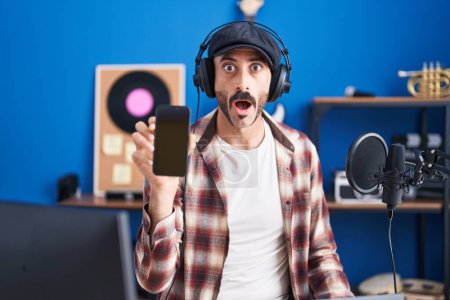 Téléchargez les photos : Homme hispanique avec barbe au studio de musique montrant smartphone effrayé et étonné avec la bouche ouverte pour la surprise, visage incrédule - en image libre de droit