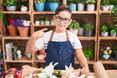 Foto de Joven niño hispano trabajando en floristería sonriendo feliz señalando con la mano y el dedo - Imagen libre de derechos