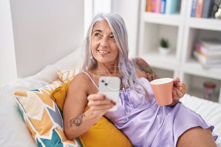 Foto de Mujer de pelo gris de mediana edad usando teléfono inteligente bebiendo café en el dormitorio - Imagen libre de derechos