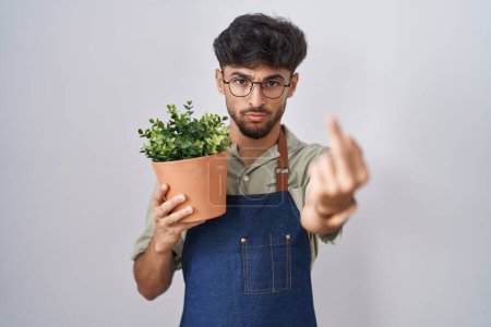 Photo for Arab man with beard holding green plant pot showing middle finger, impolite and rude fuck off expression - Royalty Free Image