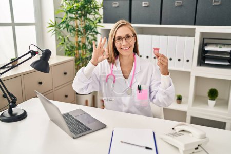 Téléchargez les photos : Jeune femme blonde médecin tenant la coupe menstruelle faire ok signe avec les doigts, sourire geste amical excellent symbole - en image libre de droit