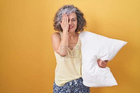 Téléchargez les photos : Femme d'âge moyen avec les cheveux gris portant un pyjama étreignant oreiller couvrant un oeil avec la main, sourire confiant sur le visage et l'émotion surprise. - en image libre de droit