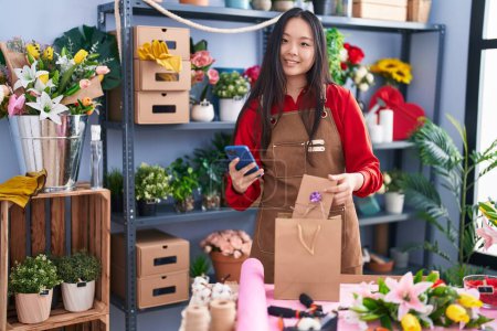 Téléchargez les photos : Jeune fleuriste chinoise utilisant un smartphone tenant un sac-cadeau à la boutique de fleurs - en image libre de droit