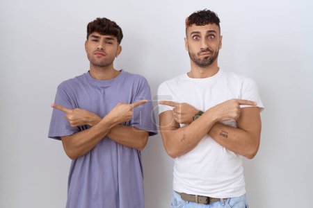 Téléchargez les photos : Couple gay homosexuel debout sur fond blanc pointant vers les deux côtés avec les doigts, direction différente en désaccord - en image libre de droit