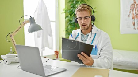 Foto de Joven caucásico hombre médico en video llamada escritura en portapapeles en clinicc - Imagen libre de derechos