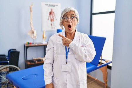 Photo for Middle age woman with grey hair working at pain recovery clinic surprised pointing with finger to the side, open mouth amazed expression. - Royalty Free Image