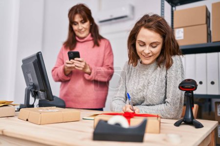 Téléchargez les photos : Deux travailleuses du commerce électronique écrivent sur un carnet à l'aide d'un téléphone intelligent au bureau - en image libre de droit