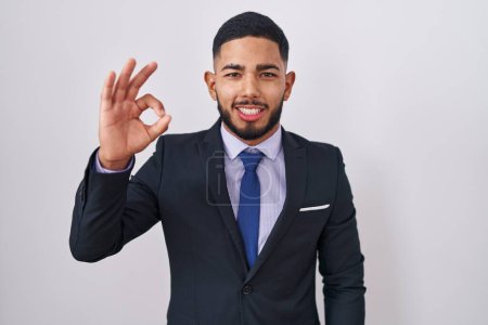 Foto de Joven hombre hispano vistiendo traje de negocios y corbata sonriente positiva haciendo bien signo con la mano y los dedos. expresión exitosa. - Imagen libre de derechos