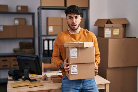 Photo for Hispanic man with beard working at small business ecommerce holding packages in shock face, looking skeptical and sarcastic, surprised with open mouth - Royalty Free Image