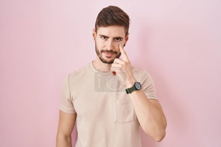 Foto de Hombre hispano con barba de pie sobre fondo rosa apuntando al ojo observándote gesto, expresión sospechosa - Imagen libre de derechos