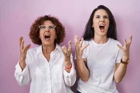 Foto de Madre e hija hispanas vistiendo una camiseta blanca casual sobre fondo rosa, locas y locas gritando y gritando con expresión agresiva y brazos levantados. concepto de frustración. - Imagen libre de derechos