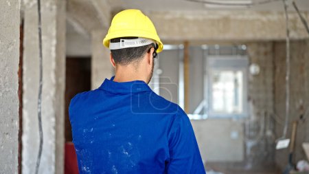 Photo for Young hispanic man worker wearing hardhat standing backwards at construction site - Royalty Free Image