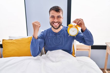 Photo for Handsome hispanic man in the bed holding alarm clock screaming proud, celebrating victory and success very excited with raised arms - Royalty Free Image