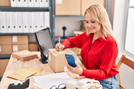 Photo for Young blonde woman ecommerce business worker writing on package using smartphone at office - Royalty Free Image