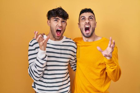 Photo for Young hispanic gay couple standing over yellow background crazy and mad shouting and yelling with aggressive expression and arms raised. frustration concept. - Royalty Free Image