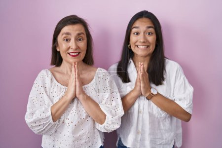 Foto de Madre e hija hispanas juntas orando con las manos juntas pidiendo perdón sonriendo confiadas. - Imagen libre de derechos